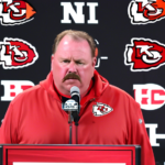 Create an image of Andy Reid, head coach of the Kansas City Chiefs, standing at a press conference podium addressing the media with concern on his face. Behind him, a large screen displays an image of