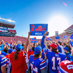 Create an image featuring enthusiastic Buffalo Bills fans celebrating outside a stadium with banners and flags, highlighting the team's 2-0 start to the 2024 season. The scene should include fans wear