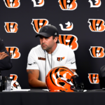 Create an image depicting an NFL press conference with Cincinnati Bengals head coach Zac Taylor looking concerned, possibly with a backdrop featuring silhouettes of wide receivers Ja'Marr Chase and Te